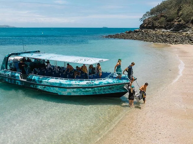 Pasajeros subiendo a un barco turístico desde una playa de arena en un día soleado.