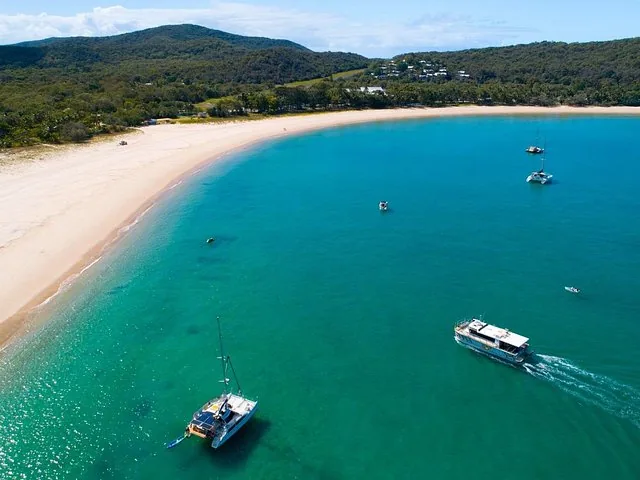 Vista aérea de una playa virgen, aguas turquesa y barcos anclados.