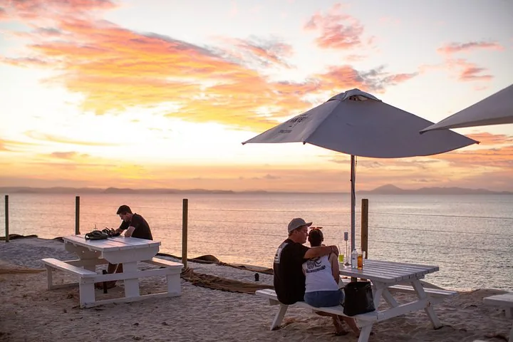 People enjoying drinks and the sunset at picnic tables on a beach.