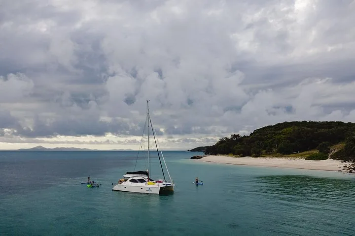Un voilier est ancré près d'une plage de sable, avec des pratiquants de paddle à proximité.